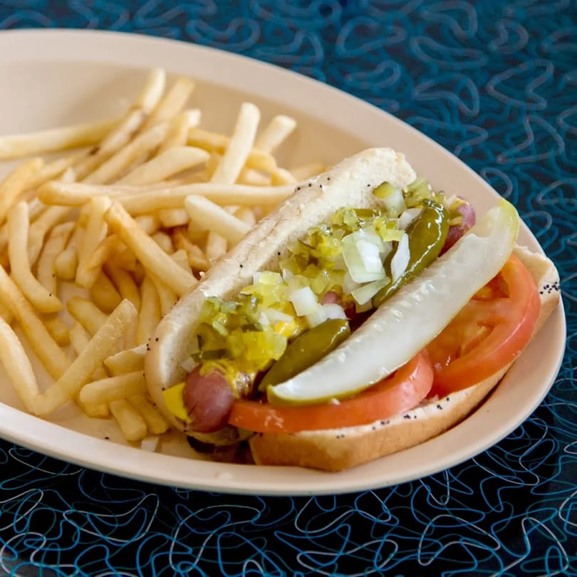 A Chicago-style hot dog with fries on a beige plate, on a blue and black patterned table.